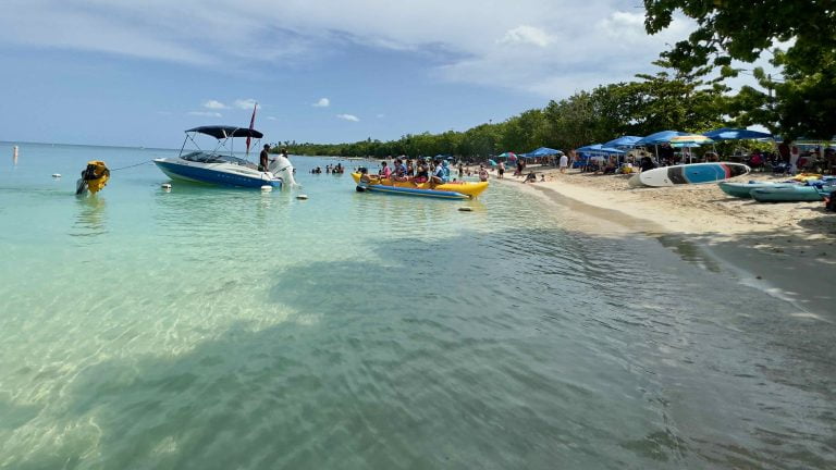 Playa Buyé de día: agua clara y poco honda, un bote cerca de la orilla y sombrillas en la arena